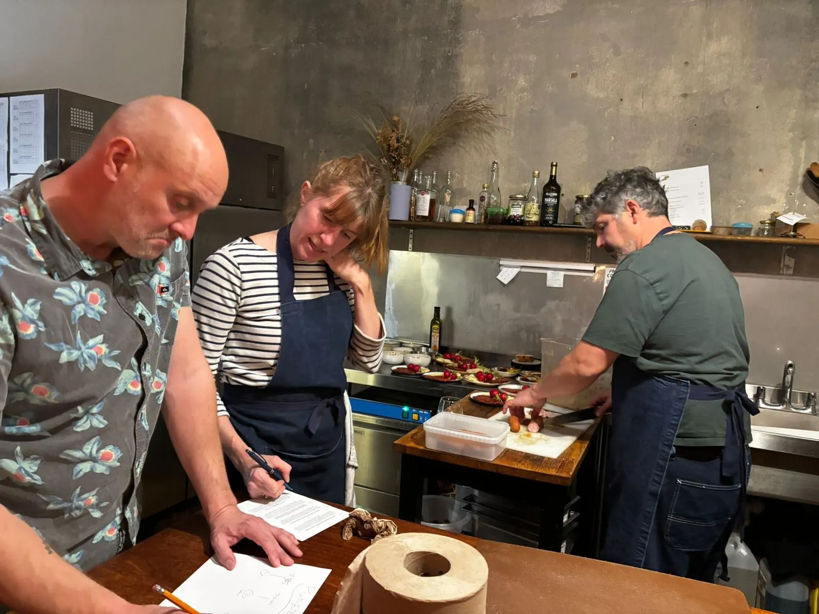 Three people in a small kitchen checking a menu sheet and slicing sausage